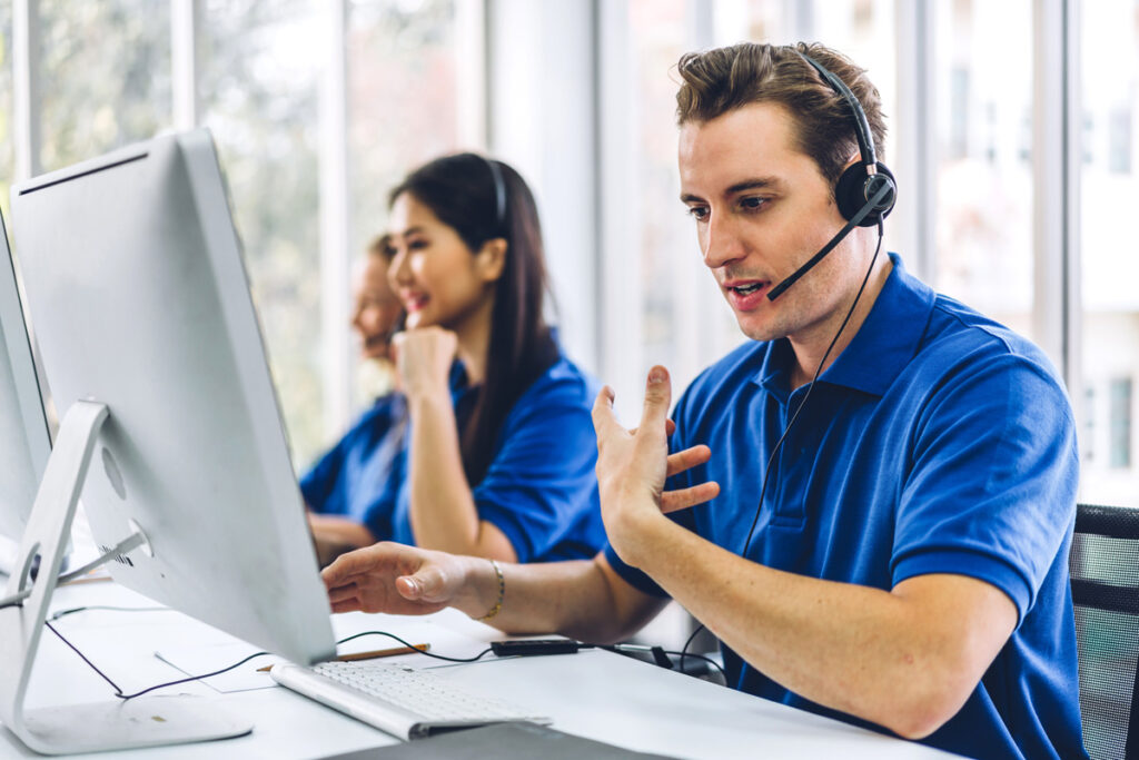 A man and his coworkers in the background, all in blue shirts, talking through headsets at a desk.